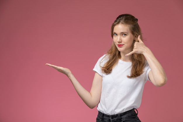 Giovane femmina attraente di vista frontale in maglietta bianca che posa con il gesto di telefonata sulla foto a colori di posa femminile del modello della parete rosa