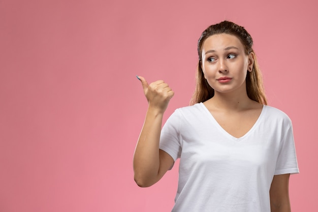 ピンクの背景にジェスチャーを指すとポーズをとって白いtシャツで正面の若い魅力的な女性