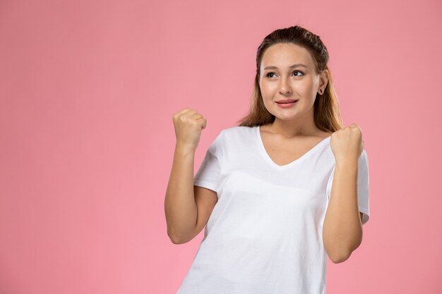 Front view young attractive female in white t-shirt posing with raised hands and delighted expression on the pink background