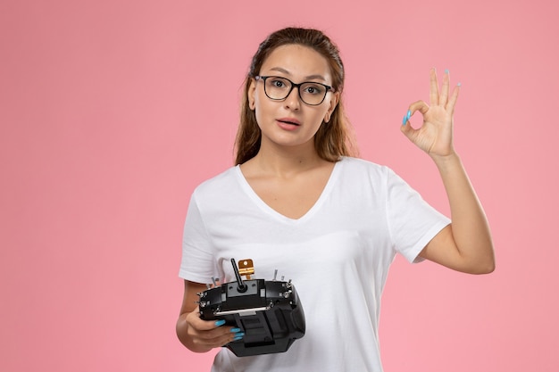 Front view young attractive female in white t-shirt posing with remote controller on the pink background