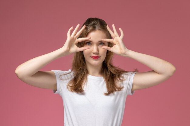 ピンクの壁モデルの女性のポーズのカラー写真でポーズをとって彼女の目を示す白いTシャツの正面図若い魅力的な女性