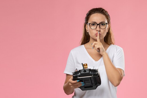 正面に沈黙の兆しを見せ、ピンクの背景にリモートコントローラーを保持している白いtシャツの若い魅力的な女性