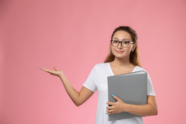 白いtシャツsmiとピンクの背景に灰色のドキュメントを保持している正面の若い魅力的な女性