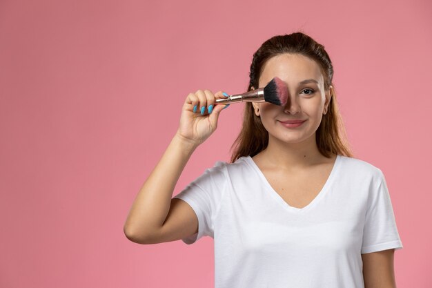 Front view young attractive female in white t-shirt smi and holding make-up brush on the pink background