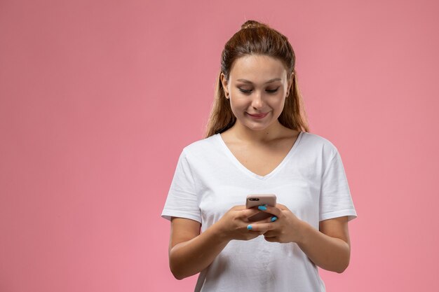 Front view young attractive female in white t-shirt smi and using a phone on the pink background