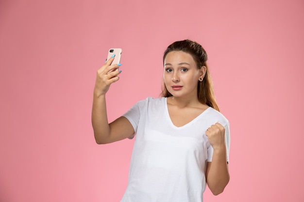 ピンクの背景に、selfieを取って白いtシャツの正面の若い魅力的な女性