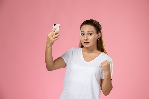 Front view young attractive female in white t-shirt taking a selfie on pink background