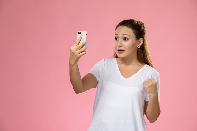 ピンクの背景に、selfieを取って白いtシャツの正面の若い魅力的な女性