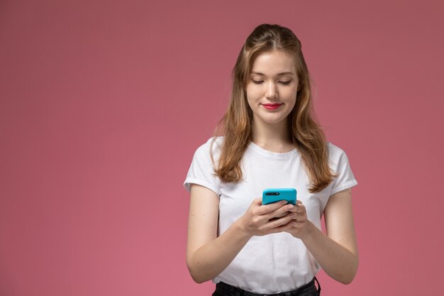 Вид спереди молодая привлекательная женщина в белой футболке, использующая телефон с легкой улыбкой на розовой стене, модель девушки позируют цветное фото