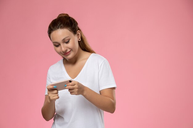 Вид спереди молодая привлекательная женщина в белой футболке смотрит видео на своем телефоне на розовом фоне
