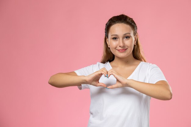 Front view young attractive female in white t-shirt with smile showing heart sign on pink background