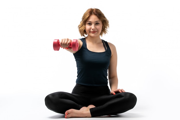 A front view young attractive girl in blue shirt and black trousers sport outfit doing workouts with dumbbells smiling on the white