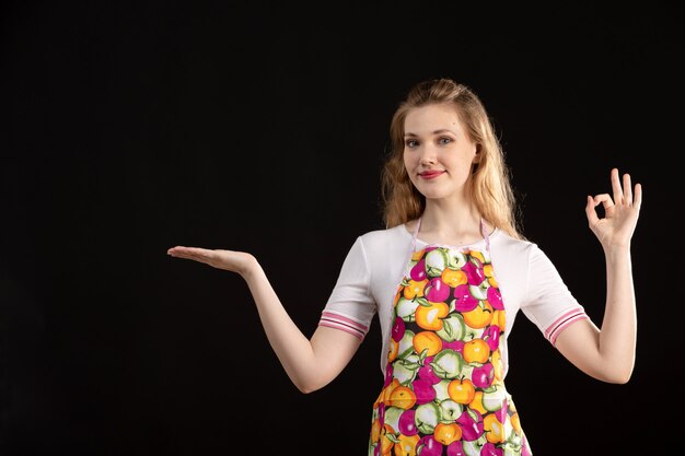 A front view young attractive girl in colorful cape showing hand signs smiling on the black background cleaning housewife