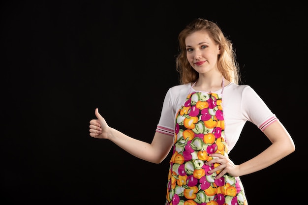 Вид спереди молодая привлекательная девушка в красочной накидке улыбается, указывая пальцами на черном фоне уборки домохозяйки