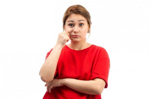 A front view young attractive girl in red t-shirt wearing black jeans pulling her cheek on the white