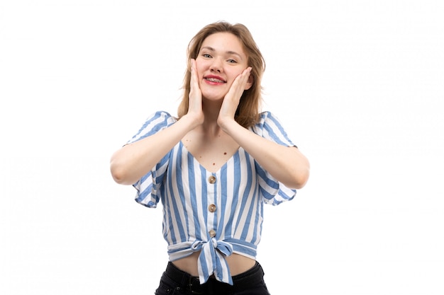 Una giovane ragazza attraente di vista frontale in maglietta bianca blu a strisce che porta i jeans neri che posano sorridere divertito sul bianco