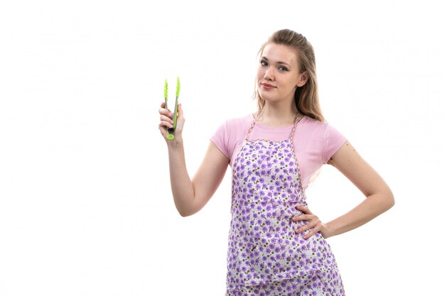 Вид спереди молодая привлекательная домохозяйка в розовой рубашке разноцветного плаща держит прибор, улыбаясь на белом фоне кухня кухня женщина