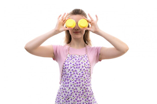 A front view young attractive housewife in pink shirt colorful cape holding yellow thing smiling on the white background cuisine kitchen female