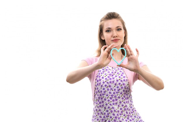 A front view young attractive housewife in pink shirt colorful cape posing smiling holding little blue heart shape on the white background cuisine kitchen female