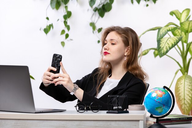 A front view young attractive lady in black jacket and white shirt in front of table working with laptop taking a selfie work business technologies