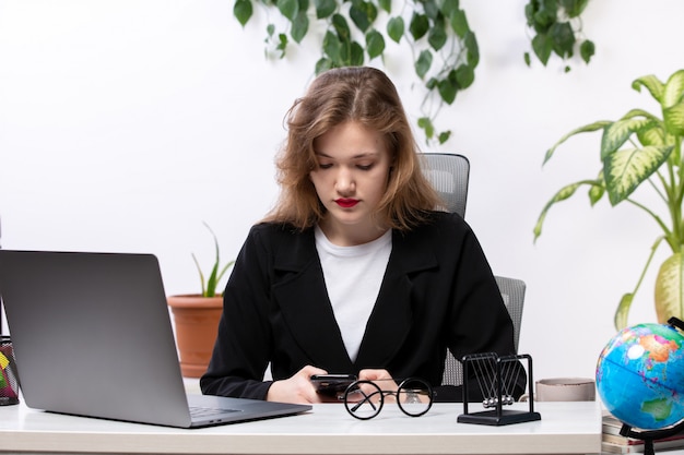 Вид спереди молодой привлекательной леди в черном пиджаке и белой рубашке перед столом, работающим с ноутбуком, используя телефонные бизнес-технологии работы