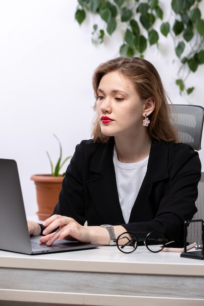 Вид спереди молодой привлекательной леди в черном пиджаке и белой рубашке перед столом, работающим с бизнес-технологиями работы ноутбука