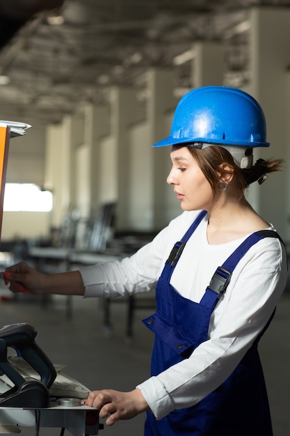 A front view young attractive lady in blue construction suit and helmet controlling machines in hangar during daytime buildings architecture construction