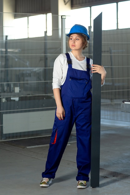 A front view young attractive lady in blue construction suit and helmet working holding heavy metallic detail during daytime looking into the distance buildings architecture construction