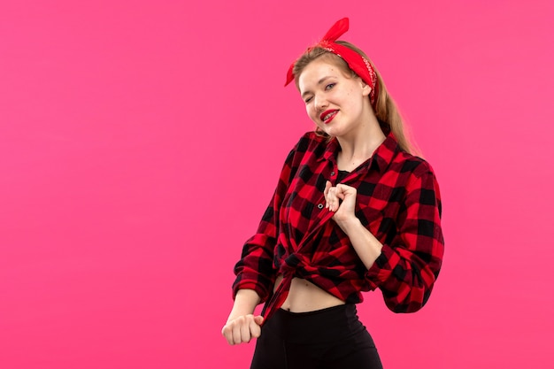 A front view young attractive lady in checkered red-black shirt black trousers smiling happy posing on the pink background young female fashion