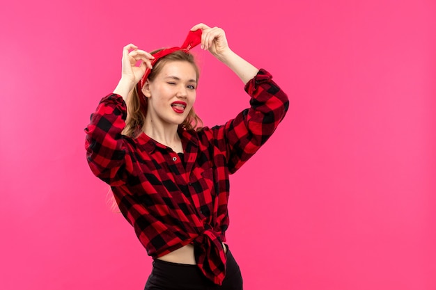 A front view young attractive lady in checkered red-black shirt black trousers smiling posing on the pink background young female fashion