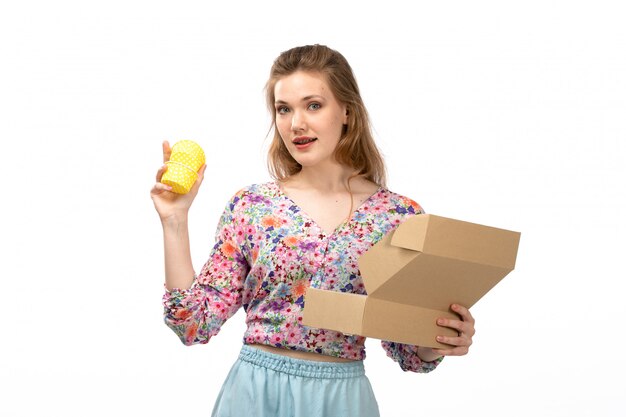 A front view young attractive lady in colorful flower designed shirt and blue skirt getting yellow present from little package on the white