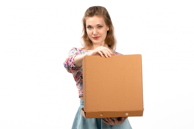 Вид спереди молодая привлекательная дама в красочном цветочном дизайне рубашки и синей юбке держит коричневую коробку на белом