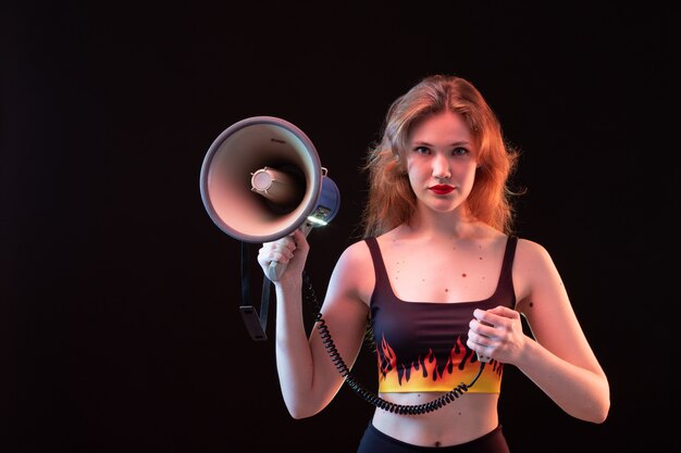 A front view young attractive lady in fire shirt and black trousers using megaphone on the black background volume loud scream