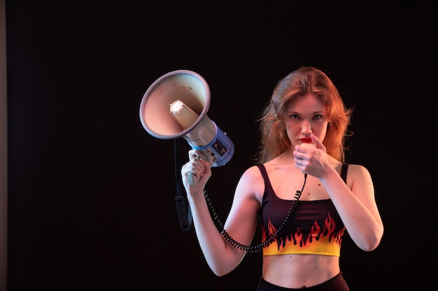 A front view young attractive lady in fire shirt and black trousers using megaphone on the black background volume loud
