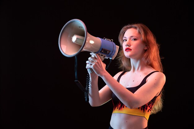 A front view young attractive lady in fire shirt and black trousers using megaphone on the black background volume loud