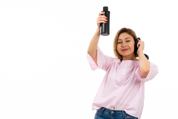 A front view young attractive lady in pink shirt and blue jeans with black earphones drinking holding black thermos black earphones smiling on the white