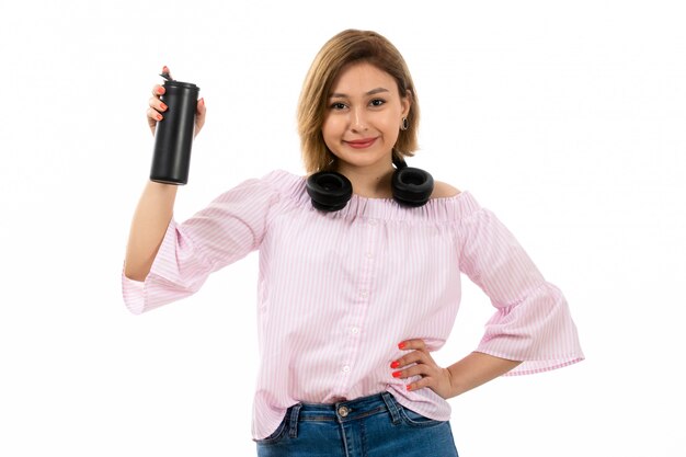 A front view young attractive lady in pink shirt and blue jeans with black earphones drinking holding black thermos smiling on the white