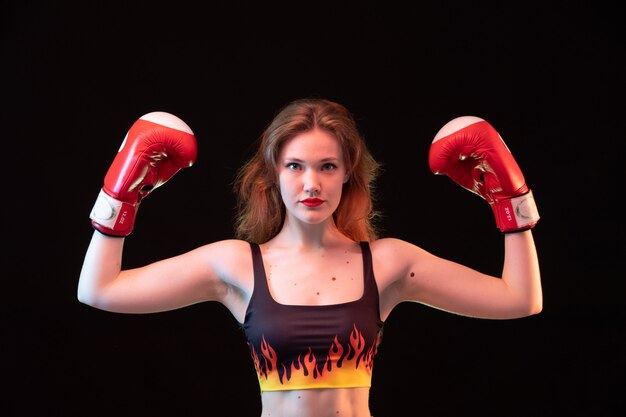 A front view young attractive lady in red boxing gloves fire shirt flexing on the black background sport boxing training