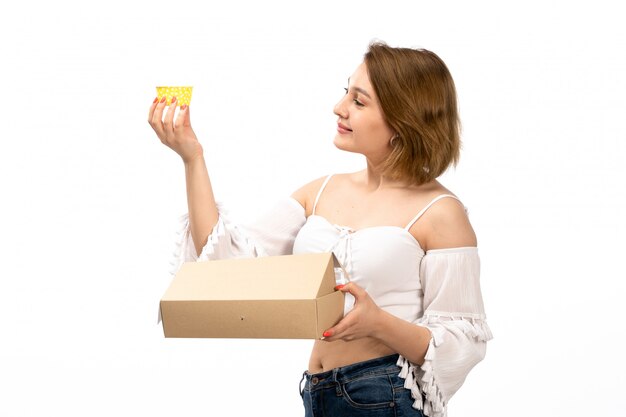 A front view young attractive lady in white shirt and blue jeans getting little package opening it on the white