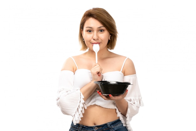 Free photo a front view young attractive lady in white shirt and blue jeans holding black bowl eating from it on the white