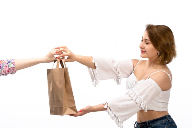 A front view young attractive lady in white shirt and blue jeans taking brown package smiling on the white