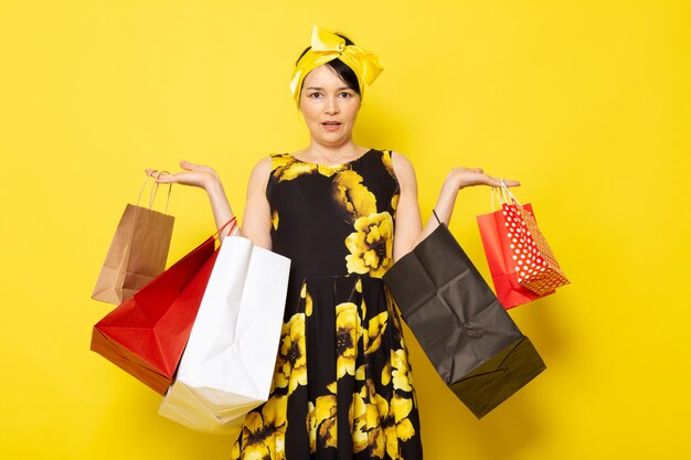 A front view young attractive lady in yellow-black flower designed dress with yellow bandage on head posing holding shopping packages on the yellow