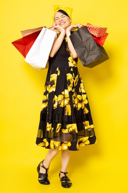A front view young attractive lady in yellow-black flower designed dress with yellow bandage on head posing holding shopping packages on the yellow
