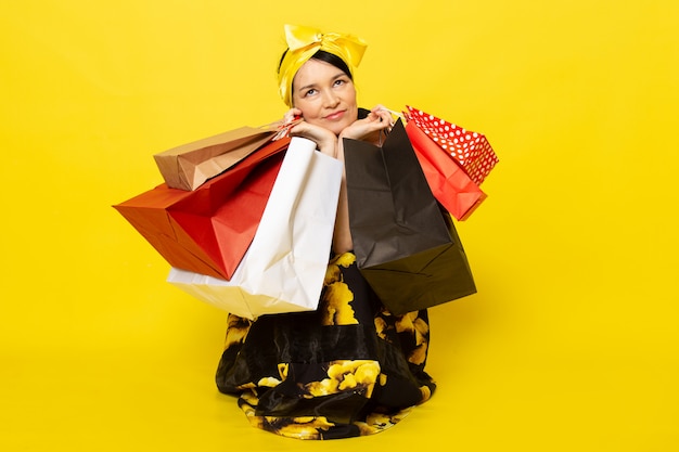 A front view young attractive lady in yellow-black flower designed dress with yellow bandage on head smiling holding shopping packages on the yellow