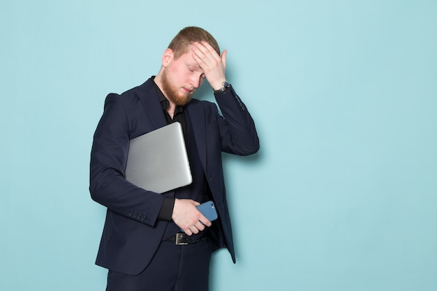 A front view young attractive male with beard in black dark classic modern suit holding grey laptop phone on the blue space