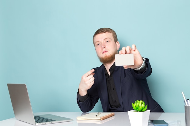 A front view young attractive male with beard in black dark classic modern suit using laptop showing white card on the blue space