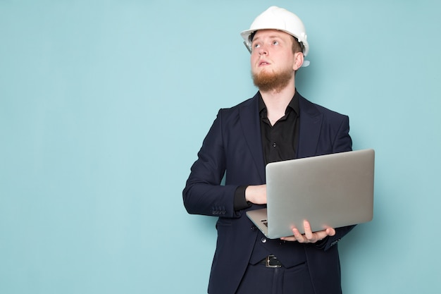 Un giovane maschio attraente di vista frontale con la barba nel casco bianco della costruzione del vestito moderno classico scuro nero che per mezzo del computer portatile grigio sullo spazio blu