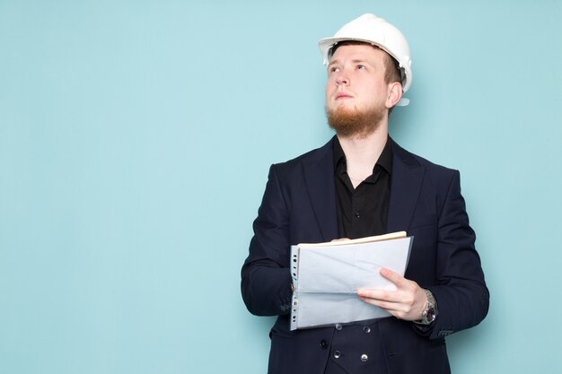 A front view young attractive male with beard in black dark classic modern suit in white construction helmet writing down notes on the blue space