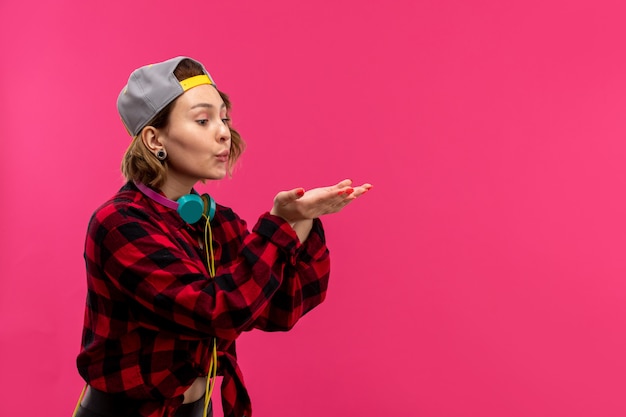 A front view young attractive woman in checkered red-black shirt black trousers with blue earphones posing sending air kisses on the pink background young female fashion