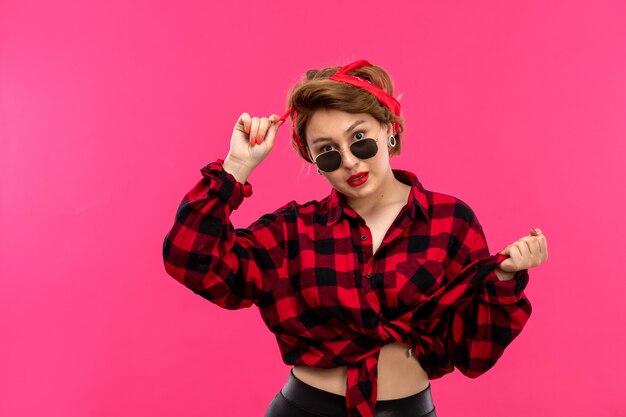 A front view young attractive woman in checkered red-black shirt black trousers with sunglasses posing on the pink background young female fashion